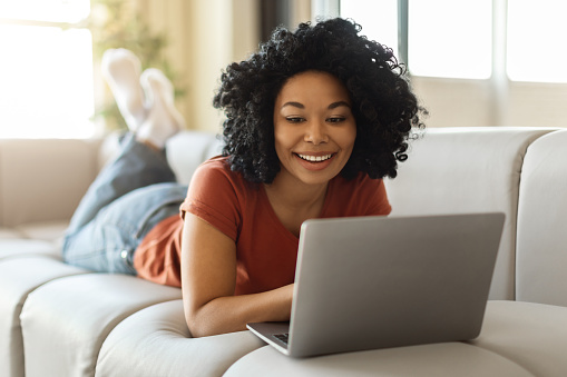 Happy Black Woman Relaxing With Laptop On Couch At Home, Smiling African American Lady Watching Movies Or Browsing Internet On Computer While Resting On Sofa In Living Room, Enjoying Weekend Pastime