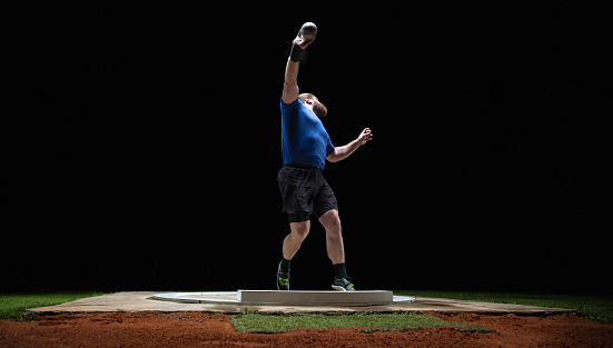 Athlete man throwing shot put ball on track during practice at night in athlete arena. Sport and healthy lifestyle concept.