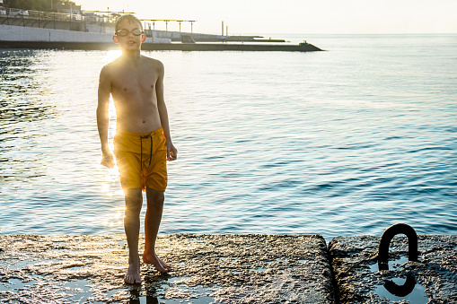 Boy with swim goggles and wet shorts stands on stone pier in cool morning. Sportive kid rests after refreshing swimming in cold sea water