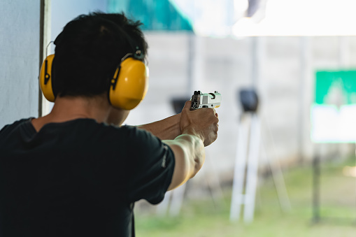 Asian shooter man wearing noise canceling headphones and black clothes practicing shooting short gun at the shooting range. Shooting sports for meditation and self-defense, recreational activities.