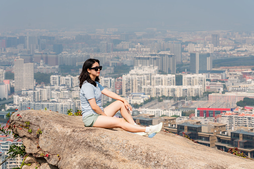 Asian female backpacker on the rock with Xiamen city skyline in the background, mountaineering, leisure and healthy living concept