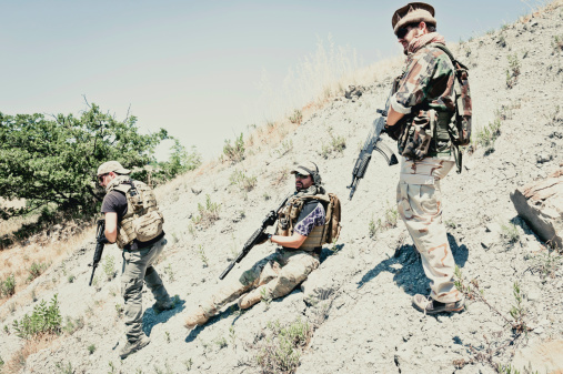 Caucasian and latin soldiers with bulletproof vest and assault rifles climbing a hill, covering each other.