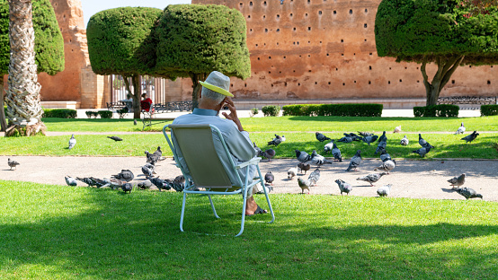 Rabat, Morocco - 9 September 2022: An old man sits on a bench in the park, surrounded by pigeons. Concept of happy retirement, simple happiness and the start of a new chapter in life
