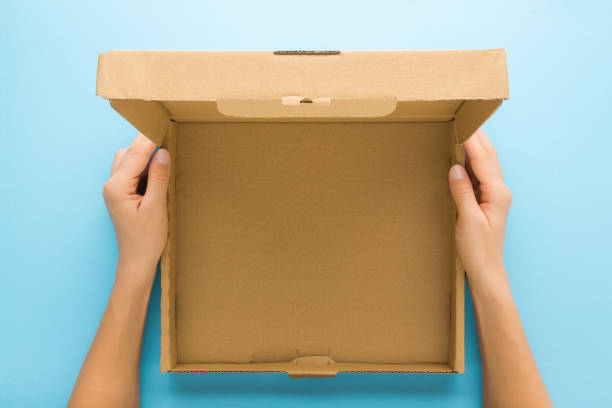 Woman hands holding opened brown carton box on light blue table background. Pastel color. Closeup. Empty place for different things, goods, food or products packaging for delivery. Top down view. manos de mujer sosteniendo una caja de cartón marrón abierta sobre fondo de mesa azul claro. color pastel. closeup. lugar vacío para diferentes cosas, mercancías, alimentos o envases de productos para su entrega. vista de arriba hacia abajo.