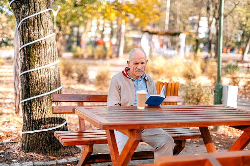 Old man spending time in city park in Autumn sitting on park bench and having coffee drink
