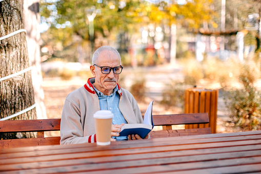 Old man spending time in city park in Autumn sitting on park bench and having coffee drink