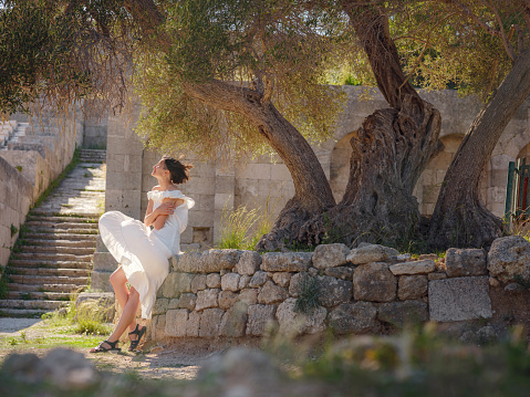 Beautiful Asian young woman in white dress outdoor. Acropolis of Rhodes Famous ruins of ancient settlement with various buildings such as stadium and theater.