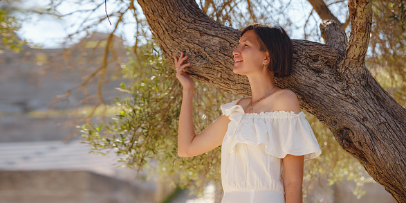 Beautiful Asian young woman in white dress outdoor near olive tree. embracing fresh air and engaging in outdoor activities. Friluftsliv concept means spending as much time outdoors as possible