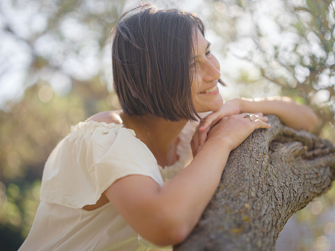 Beautiful Asian young woman in white dress outdoor near olive tree. embracing fresh air and engaging in outdoor activities. Friluftsliv concept means spending as much time outdoors as possible