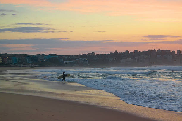 Bondi Beach - Pink Sunrise 5.59am: A surfer walks towards the sea at sunrise on Bondi Beach, Sydney. Love how there seems to be some symmetry in the shapes of the clouds and sea/sand. bondi beach sunrise stock pictures, royalty-free photos & images