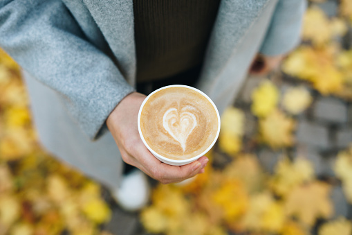 Close up of a woman's hand holding coffee to go outdoors. Yellow autumn leaves in the background. Enjoying autumn weather concept.