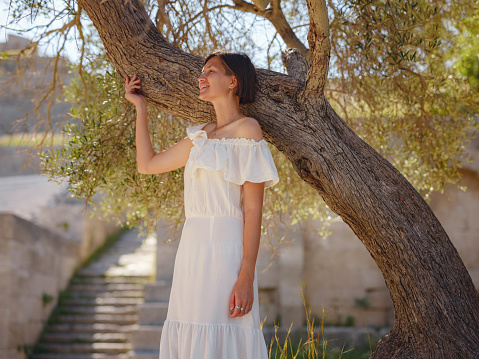 Beautiful Asian young woman in white dress outdoor near olive tree. embracing fresh air and engaging in outdoor activities. Friluftsliv concept means spending as much time outdoors as possible