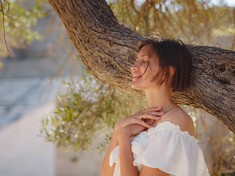 Beautiful Asian young woman in white dress outdoor near olive tree. embracing fresh air and engaging in outdoor activities. Friluftsliv concept means spending as much time outdoors as possible