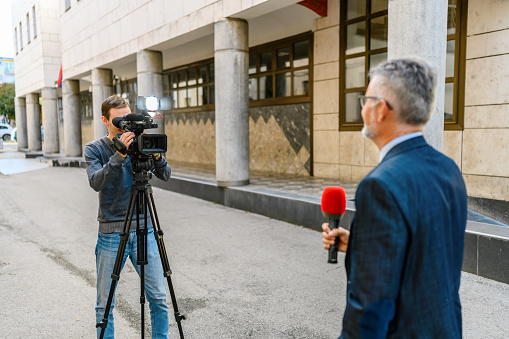 A male television reporter holding a microphone, is standing outdoors in front of a building. A male camera operator is filming him.