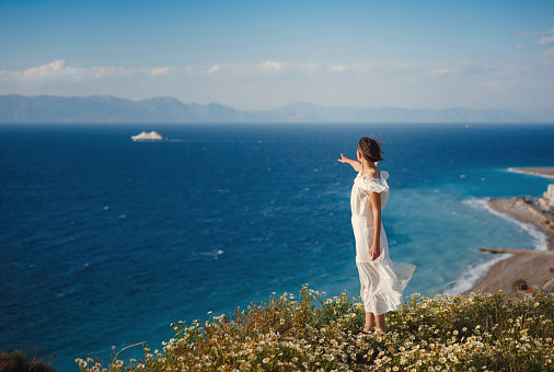 Beautiful Asian young woman in white dress outdoor in flower field under Rhodes city and above sea during sunset. embracing fresh air and engaging in outdoor activities. strong wind - element of freedom