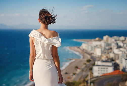 Beautiful Asian young woman in white dress outdoor in flower field under Rhodes city and above sea during sunset. embracing fresh air and engaging in outdoor activities. strong wind - element of freedom