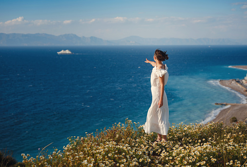 Beautiful Asian young woman in white dress outdoor in flower field under Rhodes city and above sea during sunset. embracing fresh air and engaging in outdoor activities. strong wind - element of freedom