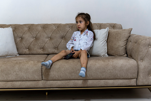 Portrait of a girl sitting on a sofa at home. Girl watching television. Girl child using television remote control.