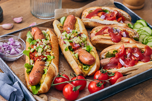 Hot dog barbecue brunch made of various Hot dog sausages or sandwiches, made with fresh organic vegetables, green salad, mustard, tomato salsa ketchup, cucumber, Spanish onion, local seasoning, herbs and spices, with French fries, served on a metal plate on a wooden rustic home or restaurant table, representing gourmet joy, a healthy protein meal, a street food lifestyle and wellbeing through healthy eating