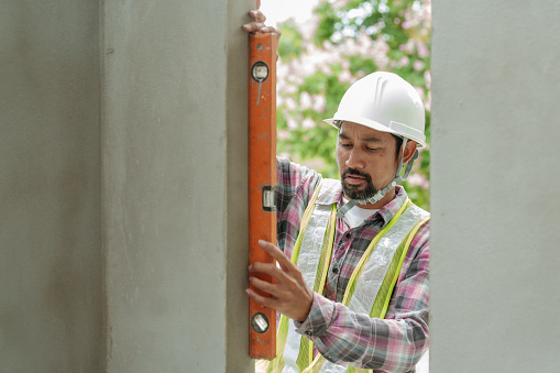 Asian man construction supervisor employee engineer Wearing white usingmeasuring stick measure straightness housing project window frame. Asian architect looks building structure diagram background.