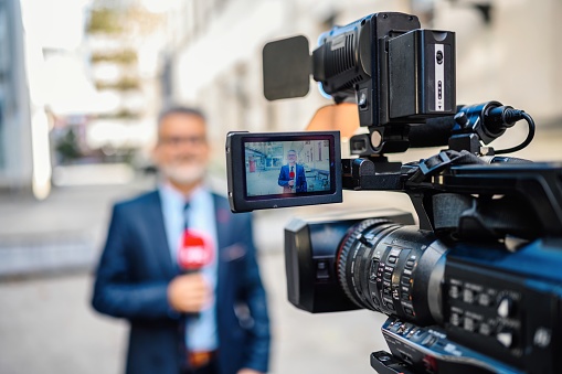 A male television reporter holding a microphone, is standing outdoors in front of a building. A male camera operator is filming him. Selective focus.