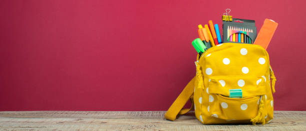 istock Yellow backpack with white polka dots with different colorful stationery on table. Burgundy background. Back to school. Banner design. 1776547294