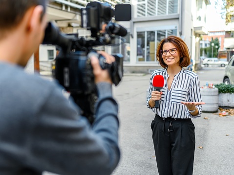 Female television reporter holding a microphone, she is standing outdoors in front of a building. Male camera operator is filming her.