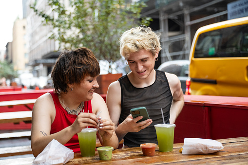 Waist up view of young New Yorkers sitting together at sidewalk cafe table with drinks and snacks, smiling as they check content on portable device.