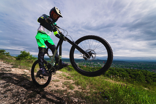 an athlete performs a stunt while standing on a wheelie on a rock, overlooking green forests and mountains. cycling extreme sport, man in full protection of the whole body and head