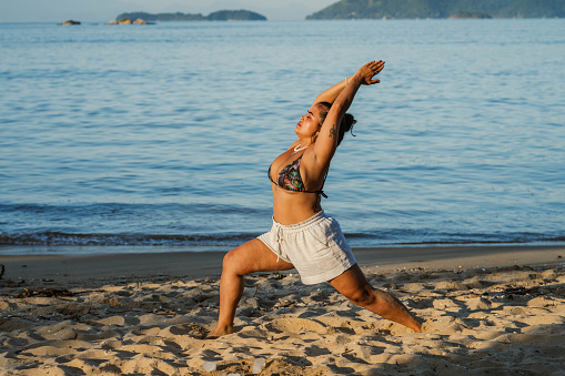 Young woman in swimwear standing in the crescent lunge pose while practicing yoga on a sandy beach in summer