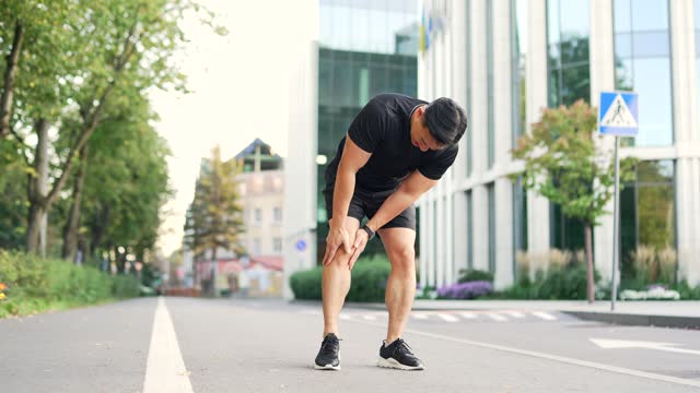Asian runner athlete with muscle pain in city street. Man massaging stretching, trauma injury while jogging outdoors in park.