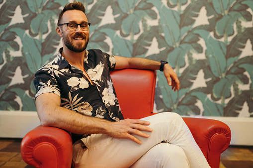 Portrait of a smiling man wearing glasses and a floral-patterned shirt sitting in a read leather chair