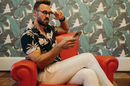 Stylish man wearing glasses and a floral-patterned shirt reading a text message on his mobile phone while sitting in a read leather chair