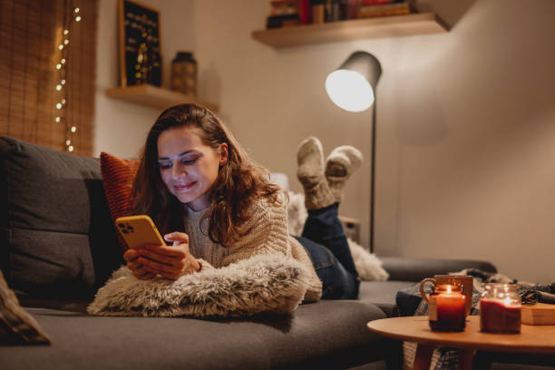 A young woman spends a cozy winter evening at home lying on the sofa looking at her smartphone. Winter holidays, Christmas and online surfing concept stock photo