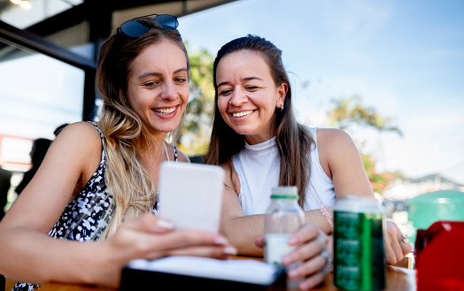 Two female friends looking at something on a mobile phone while sitting together at a cafe table outdoors
