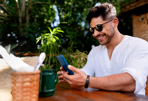 Smiling young man wearing sunglasses looking at his cell phone while sitting at outdoor coffee shop