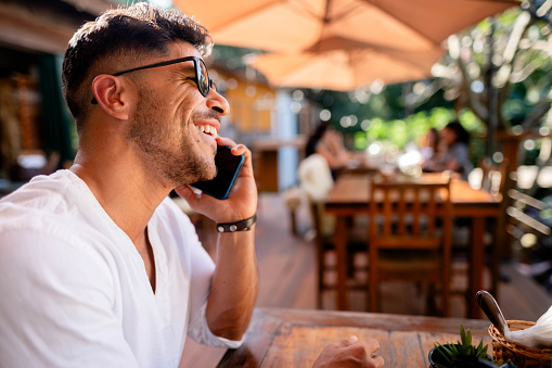 Side view of handsome young man sitting at restaurant table talking on his mobile phone and smiling