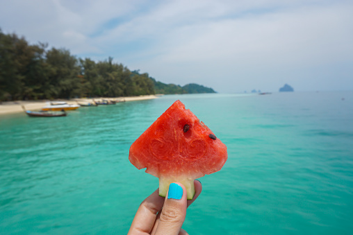 Red piece of watermelon in hand against the background of the blue sea. Cropped Hand Holding Watermelon Against Blue Ocean.