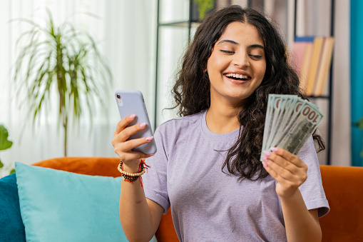 Planning budget. Rich happy Indian woman use smartphone calculator app count money cash, calculate domestic bills at home. Girl satisfied of income earnings win saves money for planned vacation gifts