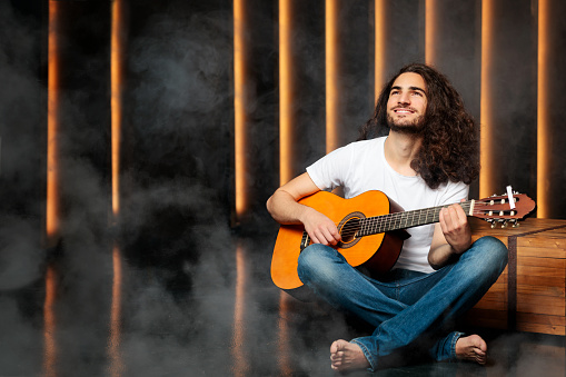 Talented Arabic Young Man With Guitar Composing New Song Sitting On Floor And Playing Chords In Dark Studio With Smoke, Looking Away Dreaming About Successful Musical Career. Copy Space