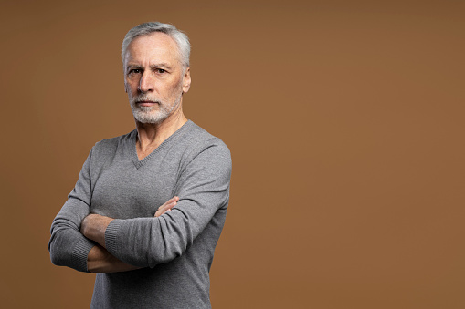 Portrait of grey haired serious senior man with arms crossed isolated on brown background. Successful business
