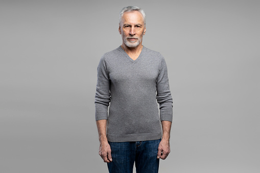 Confident elderly gray-haired bearded man 60s years old, wearing gray long sleeve and casual denim jeans, looking confidently at camera, isolated on grey background. People emotions lifestyle concept.