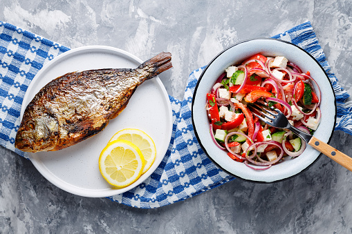 Whole baked dorado fish with lemon and vegetable salad feta cheese on gray background. Top view
