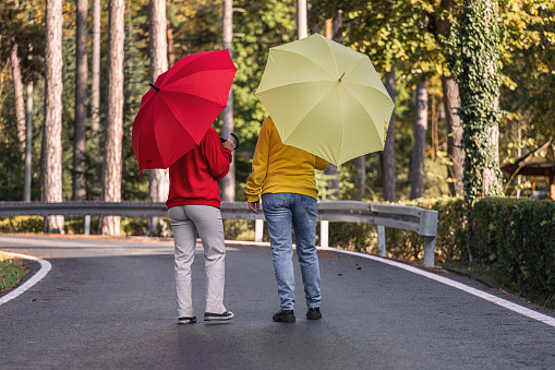 Back view of two women enjoying an autumn forest, they wearing red and yellow hoodies and holding umbrellas