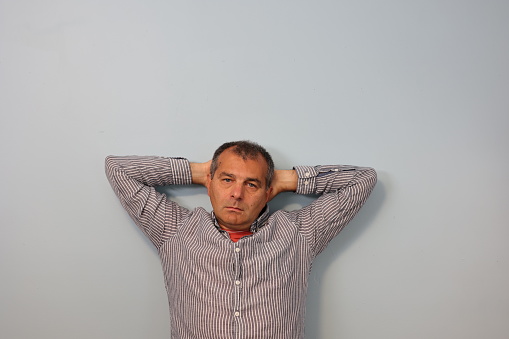 Mature male looking confused leaning on gray wall with his hands up