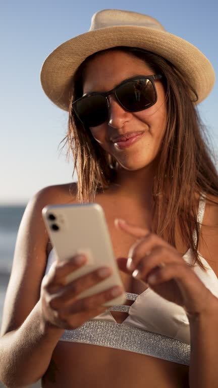 portrait young adult smiling latin american woman holding and looking a cellphone on the beach