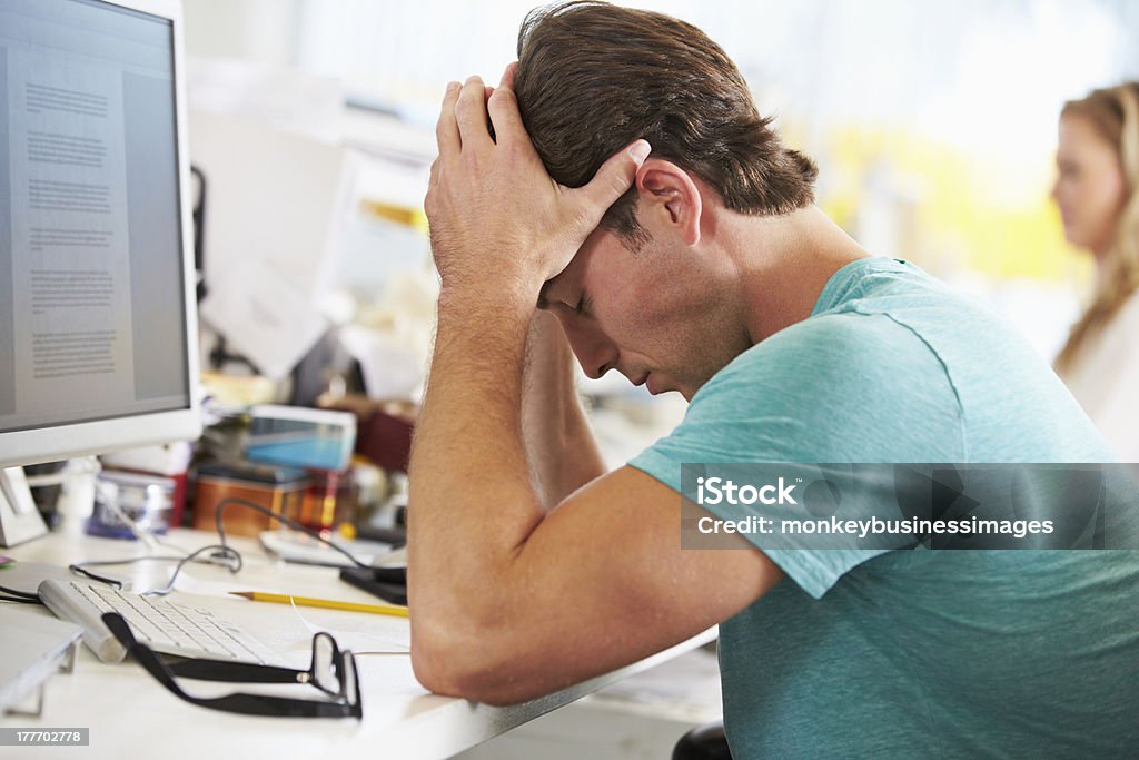 Stressed Man Working At Desk In Busy Creative Office Stressed Man Working At Desk In Busy Creative Office Stressed Graphic Design Studio Stock Photo