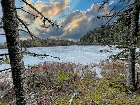 Panoramic view of snow-covered lake in northern Sweden at sunrise. From the foreground, the newly fallen snow has formed a perfectly smooth white surface stretching all the way to the other side of the lake where the backlit trees in a pine forest are silhouetted against a blue sky with low clouds. To the left, the rising sun in the middle of the lake, part of 