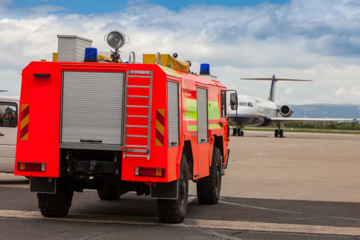 Red airport fire truck driving on the tarmac in front of the incoming airplane