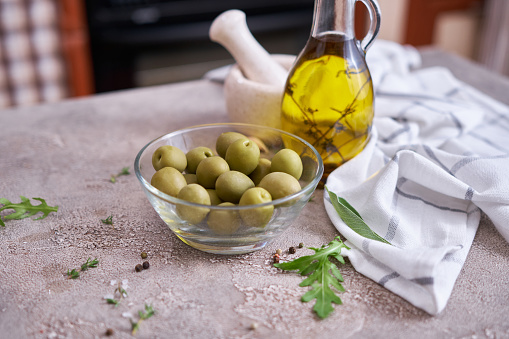 Canned green olives in a glass bowl and small bottle of olive oil with herbs at domestic kitchen.